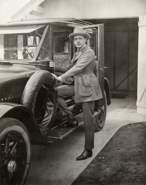 Silent film actor Niles Welch is stepping up onto the running board of a 1918 Hudson Super Six touring limousine. He wears a casual suit with a belted coat and a soft, wooly homburg hat.

Original caption:
"For recreation Niles Welch feels convinced there's nothing like an automobile, especially if you run it yourself on those matchless Southern California roads."
