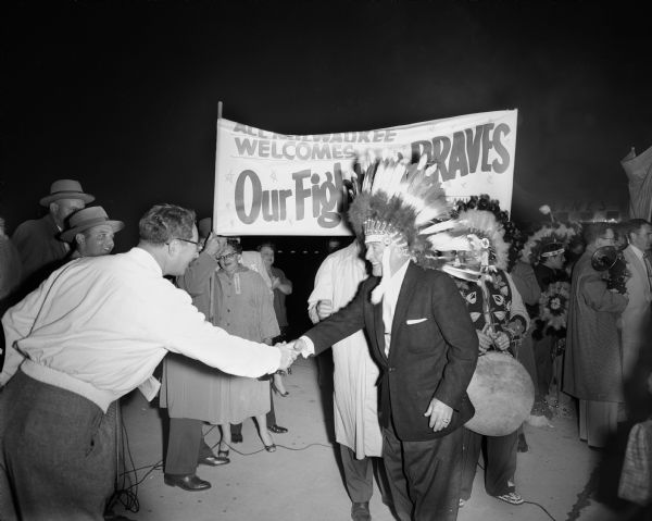 Original caption from the <i>Milwaukee Journal</i>: "Manager Fred Haney, wearing an Indian headdress, reached out to shake the hand of a greeter at Gen. Mitchell field at the Braves' homecoming Sunday night. The war bonnet was placed on him soon after he left the plane."  The rally was to welcome the Milwaukee Braves home from St. Louis, where the team had lost two of its final three games and the 1956 National League pennant.