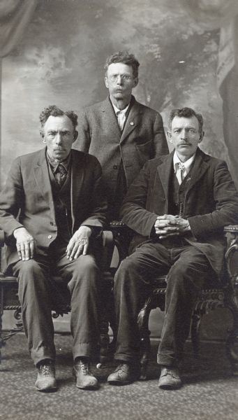 Full-length studio portrait of three men, two sitting and one standing in front of a painted backdrop.