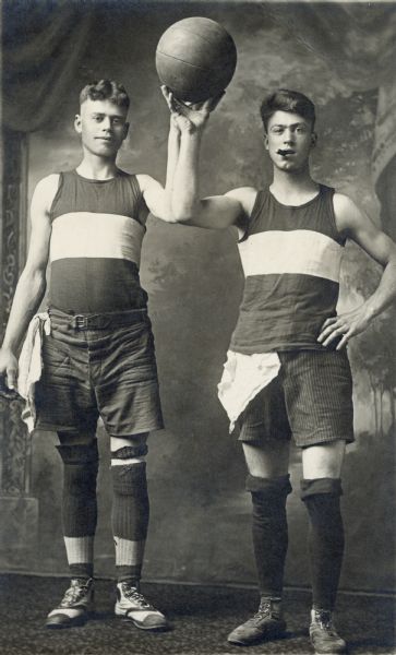 Full-length studio portrait of Rudolph Faust and his brother jointly holding up a basketball in front of a painted backdrop. The Faust brother on the right has a cigar in his mouth.