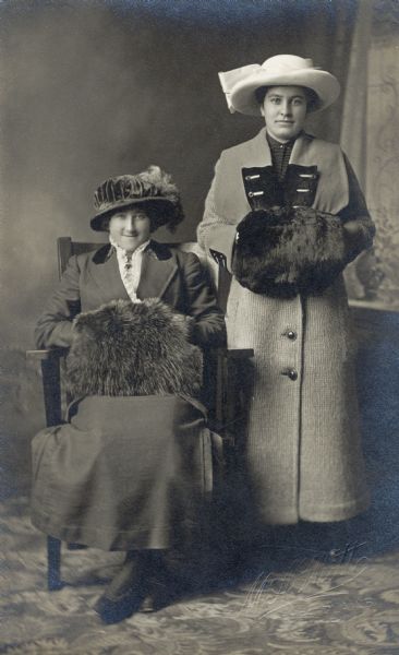 Full-length studio portrait of two women, Mrs. John Zander (Mamie Endres Zander), and Catherine Endres Mick, wearing fashionable clothing, including muffs, coats, dresses and hats. They are posing in front of a painted backdrop.