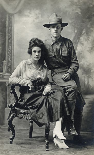 Full-length studio portrait of a woman seated in a chair, and a soldier dressed in a First World War uniform sitting on the arm of the chair. They are posing in front of a painted backdrop.
