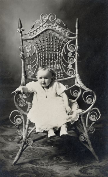 Studio portrait of an infant dressed in white sitting on a large, wicker chair.