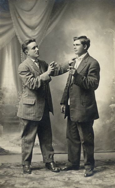 Full-length studio portrait of two men in front of a painted backdrop. The men are dressed in business suits, holding cigars in their hands and using hand gestures to emulate conversation.