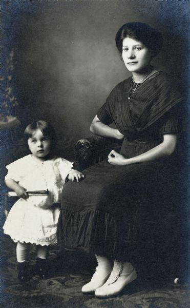 Studio portrait of mother and child in front of a painted backdrop. The woman is seated and wearing a black dress, the toddler is standing and dressed in white.
