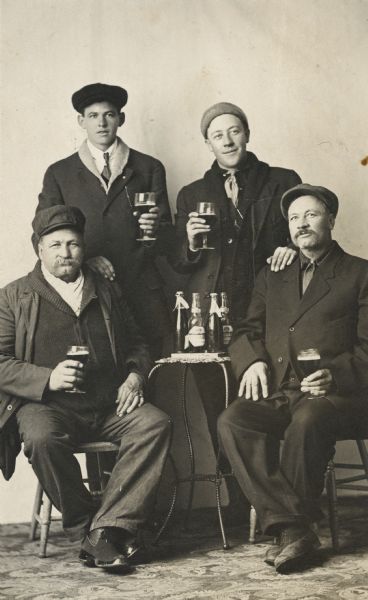 Studio portrait of two fathers and their sons holding drinks in front of a painted backdrop. In the front row, from left to right, are Jacob Birrenkott of Cross Plains, and Peter Birrenkott of South Dakota. In the back row, from left to right, are John H. Birrenkott of Cross Plains, and John Birrenkott of South Dakota.