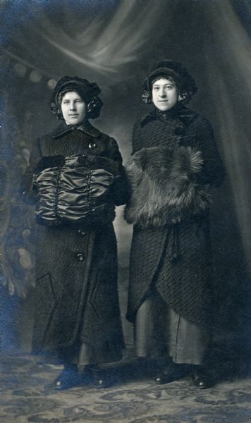 Full-length studio portrait of two women standing in front of a painted backdrop. They are dressed in black and are wearing fashionable coats, muffs and hats.