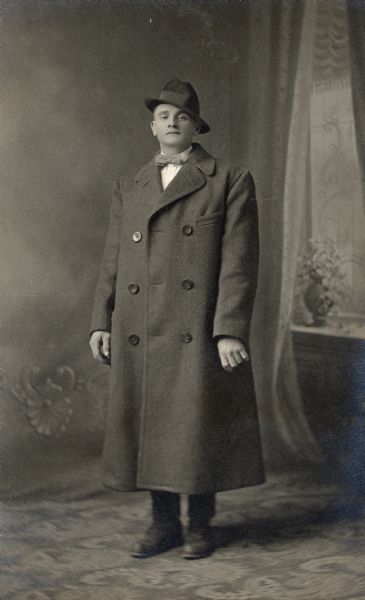 Full-length studio portrait of a man wearing a long overcoat and a bow tie in front of a painted backdrop. His hat is tilted at a jaunty angle.