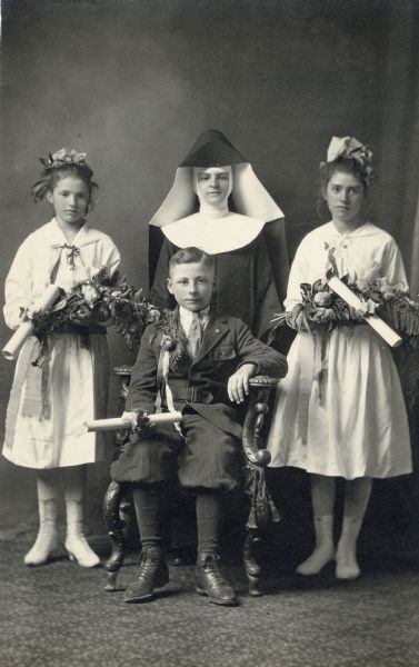 Studio portrait of three children with flower bouquets in front of a painted backdrop. One boy is seated and two girls are standing. All of the children are holding a scroll, probably a graduation diploma. A nun stands in between the two young girls.