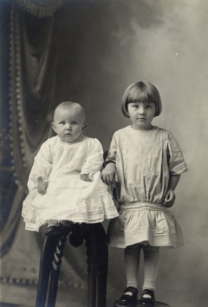 Studio portrait of two children in front of a painted backdrop. The infant is sitting on a stool, and a young girl is standing on a platform.