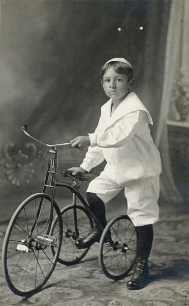 Full-length studio portrait in front of a painted backdrop of a young boy with a tricycle.