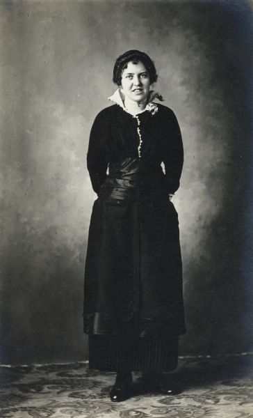 Full-length studio portrait of a woman in front of a painted backdrop. She is dressed in black and has her hands behind her back.