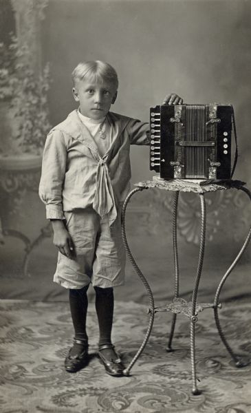 Full-length studio portrait of boy in front of a painted backdrop. He is standing with his arm resting on an accordion placed on a table.