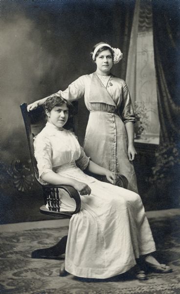 Studio portrait of two women in front of a painted backdrop. One woman is seated and one is standing, are they are dressed in fashionable dresses. The woman standing is wearing a headband in her hair.