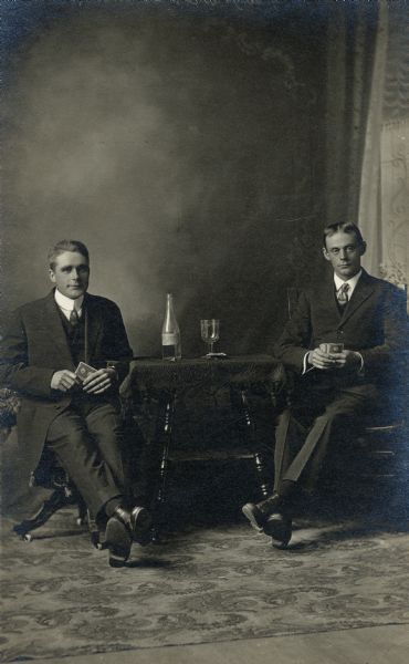 Studio portrait in front of a painted backdrop of two men sitting at a table playing cards. On the table is a bottle of wine and a glass. The man on the left is unidentified, but the man on the right is Will Fisher of Waukesha.