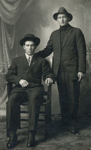 Studio portrait in front of a painted backdrop of two well-dressed men, Karl Faust, who is sitting, and Mike Endres, standing. They are wearing suits and hats and are both smoking cigars.