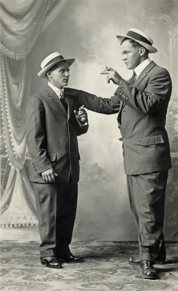 Studio portrait of two men dressed in business suits wearing hats and holding cigars in front of a painted backdrop. Using hand gestures, a suggested notion of conversation is captured.