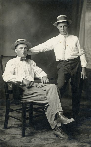 Studio portrait in front of a painted backdrop of two men smoking cigars and wearing summer hats. Joseph Bowar is sitting, and his brother, Jacob Bowar, is standing next to him.