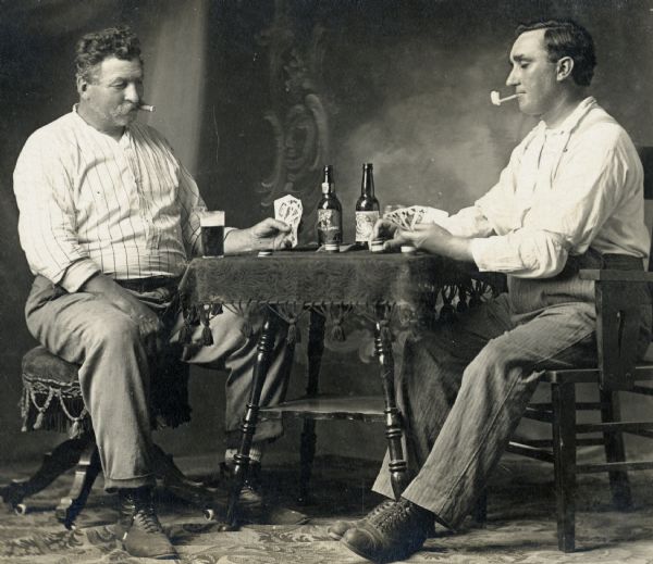 Studio portrait of two men in front of a painted backdrop playing poker while smoking and drinking beer. The man on the left is Jacob Birrenkott, and the man on the right is unidentified.