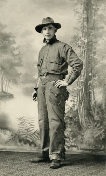 Studio portrait in front of a painted backdrop of Mike Virnig, standing with a cigarette in his hand. He is dressed in western style clothing, possibly for riding horses.