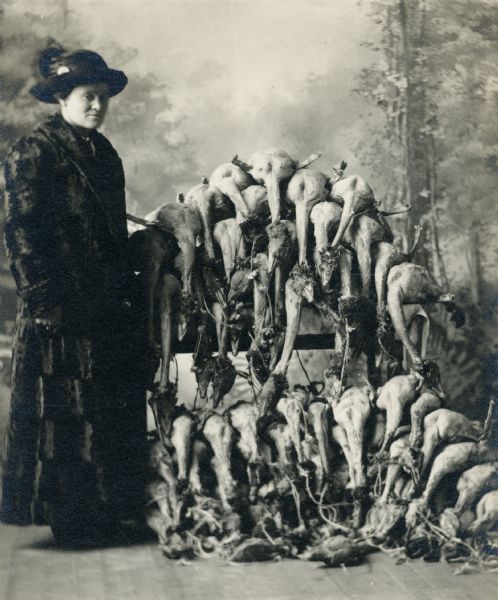 Studio portrait in front of a painted backdrop of a woman dressed in a fur coat and wearing a hat standing next to a display of defeathered geese.