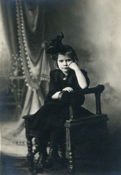 Studio portrait in front of a painted backdrop of Eleanor Faust Fuller, dressed in black with a black bow in her hair.