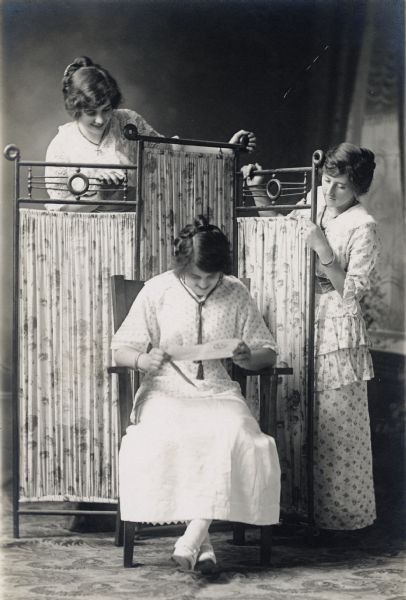 Studio portrait in front of a painted backdrop of three women posing around a folding screen. From left to right, Mamie Meyerkiester, Gertrude Boehnan Endres, and Mamie Endres Zander. Gertrude is reading a piece of paper she is holding in her hands.