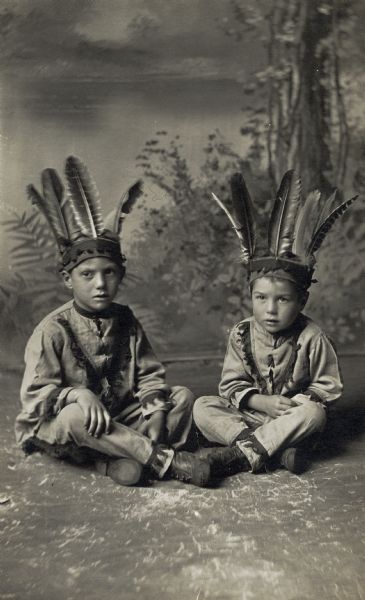 Studio portrait in front of a painted backdrop of two boys, Bill and Hubby Schmity, sitting with legs crossed on the floor and dressed in Indian costumes.