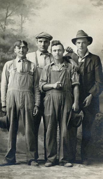 Full-length studio portrait in front of a painted backdrop of four men, all dressed in overalls, standing together. One man is holding a cigar. The two men in the back row are wearing their hats, and the two men in front are holding their hats.