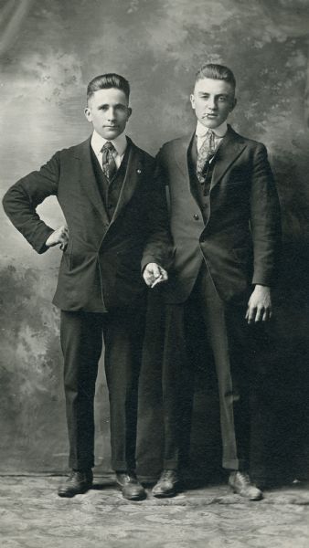 Full-length studio portrait of two unidentified men in front of a painted backdrop standing for their studio portrait. They are both dressed in suits and ties and smoking cigarettes.