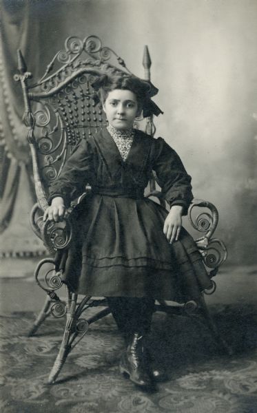 Studio portrait in front of a painted backdrop of young girl wearing a dark dress with her hair pulled back in fashionable bows. She is sitting on an ornate wicker chair.