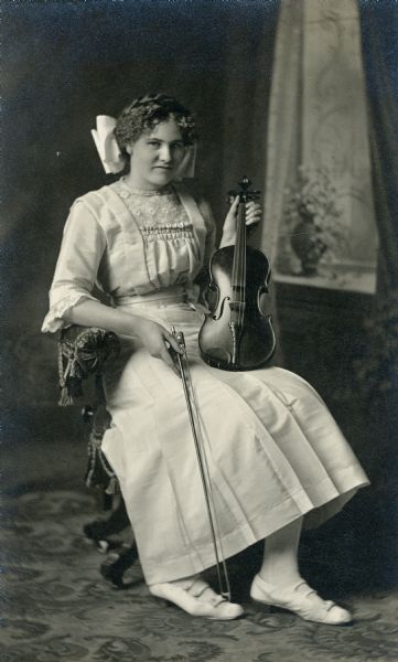Studio portrait in front of a painted backdrop of a woman sitting in a chair holding a violin on her knee. She is holding the bow in her other hand. She wearing a large white bow in her hair.