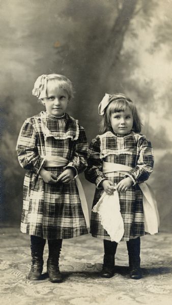 Studio portrait in front of a painted backdrop of two young girls, both dressed in plaid dresses and white bows in their hair.