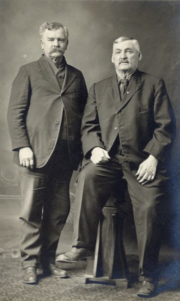 Studio portrait in front of a painted backdrop of two elderly men in suits, one standing and the other, James Gorman, sitting on a table.
