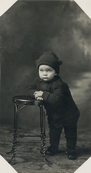 Studio portrait of a toddler leaning against a metal stool, dressed in a winter coat and hat in front of a painted backdrop.