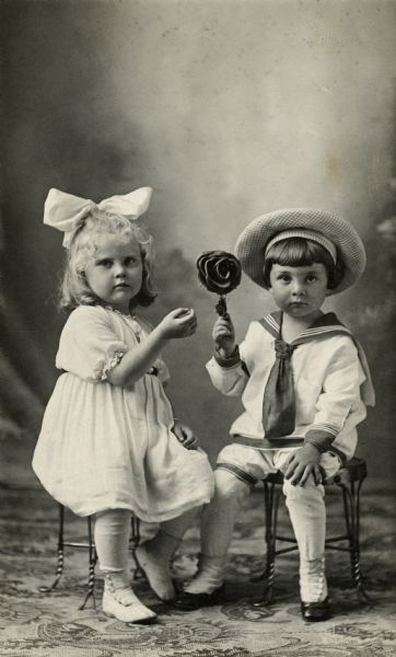 Studio portrait of two children, Liane Faust Hilgers and Harold Pick, sitting on stools in front of a painted backdrop. Harold is holding up a rose and Liane is gesturing to it. Both children are wearing fashionable clothing and Liane has a large white bow in her hair.