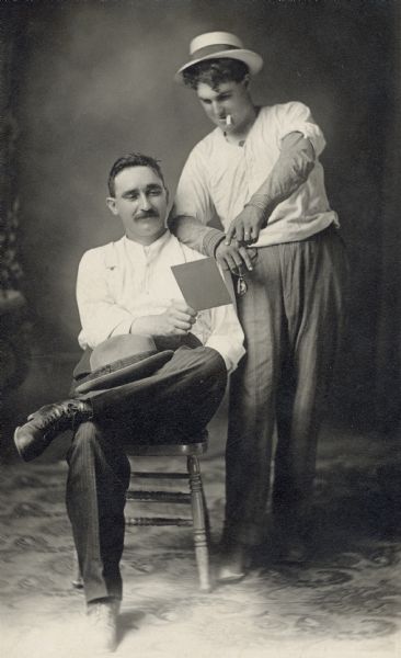Studio portrait in front of a painted backdrop of two men looking at document that Mel Esser, sitting, is holding in his hand. Charlie "Breaky" Brendler, wearing a hat and smoking a cigarette, is standing next to him.