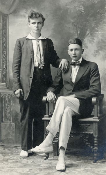 Two young men, Tom Dalton, standing, and Jerry Kraemer, sitting in front of a painted backdrop. Both are wearing suits and ties for their studio portrait.