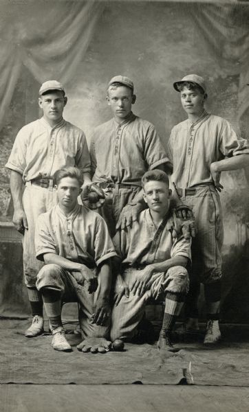 Studio portrait of six members of the baseball team posing in front of a painted backdrop. Left to right, seated: Nick Marking, Peter Burke. Standing: Matt Pick, August Eller, and Rudolph Faust.