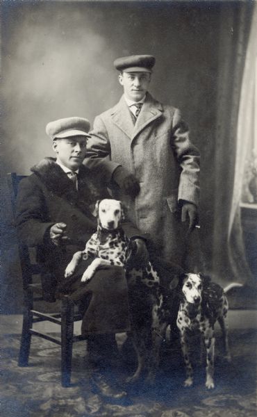 Studio portrait in front of a painted backdrop of two men wearing hats and coats, holding cigars and posing with two Dalmation dogs.