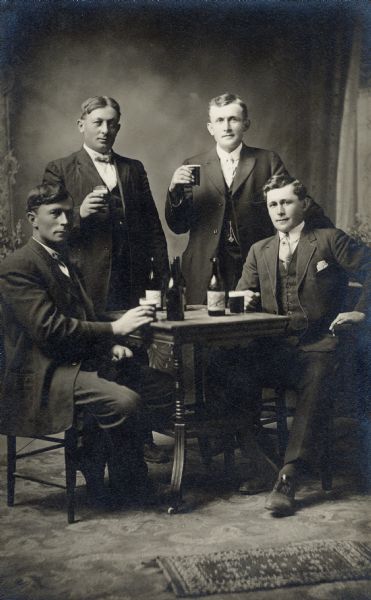 Studio portrait of four well-dressed men posed around a table drinking beer in front of a painted backdrop.