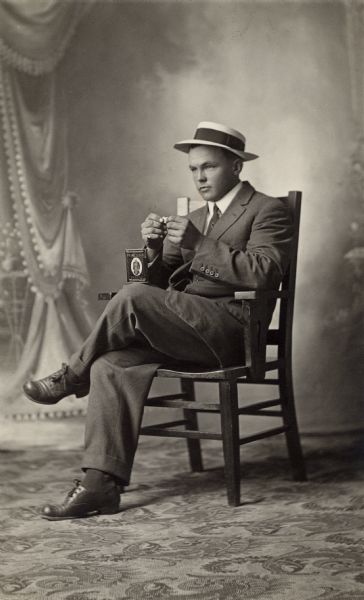 Studio portrait of a man sitting in a chair rolling a cigarette in front of a painted backdrop. A tin of Prince Albert Tobacco sits on his knee.