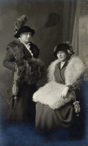 Studio portrait of two women wearing fashionable fur muffs, shawls and hats posing in front of a painted backdrop. On the left is Angeline Saeman Kalschuer, and seated is Catherine Karch Saeman. Both women also have decorative purses hanging from their wrists.