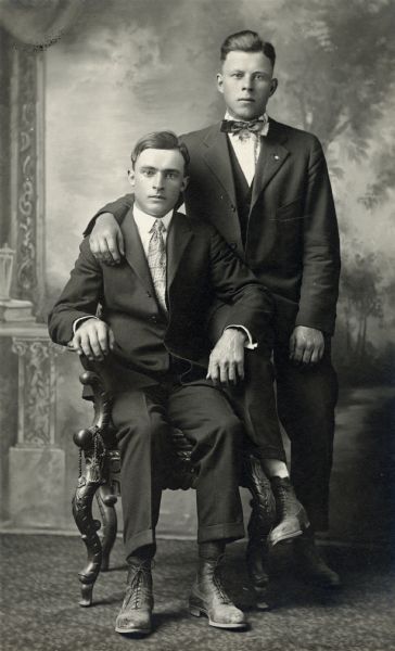 Studio portrait of two men, an unidentified man sitting in a chair, and Leo Meier, who is standing and resting his hand on the other man's shoulder. Each man is well-dressed in a suit and tie and are posing in front of a painted backdrop.