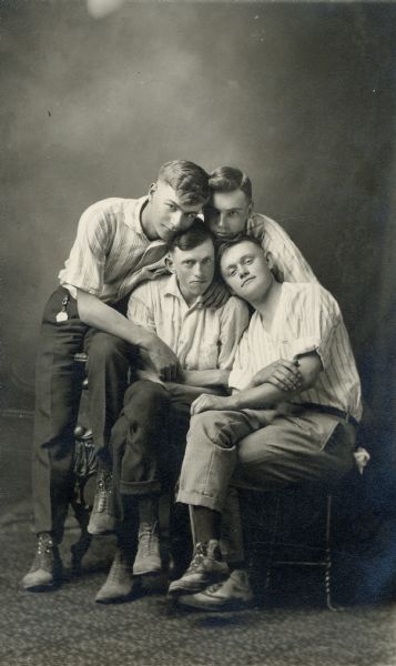 Four young men pose in front of a painted backdrop with their heads together for a group studio portrait.