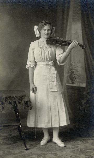Studio portrait in front of a painted backdrop of woman posing with her violin and bow. She wears a large white bow in her hair.