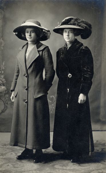 Studio portrait in front of a painted backdrop of two women wearing fashionable long coats and large hats with hatpins.