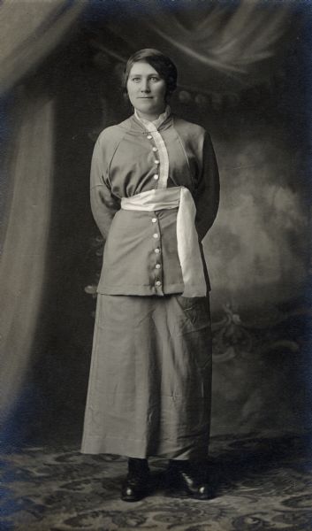 Studio portrait in front of a painted backdrop of Nellie Faust, who is wearing a dress with a large sash belt.