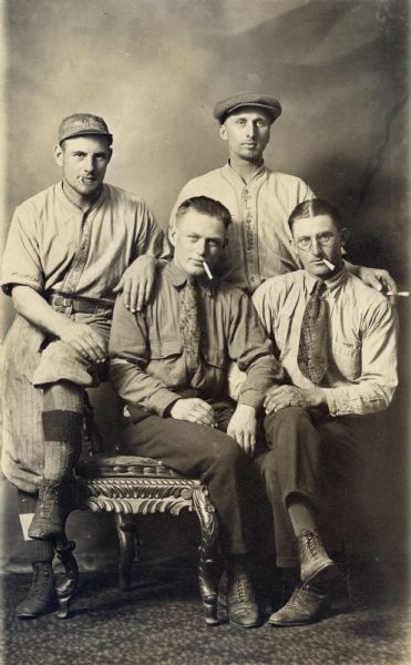Studio portrait in front of a painted backdrop of four men with cigarettes. From left to right, Mel Pick, Charlie Lenter of Milwaukee, Alphouse Frank, and Frank Schulenburg. Mel and Alphouse are wearing Cross Plains baseball uniforms, while Charlie and Frank are wearing a shirt and tie.