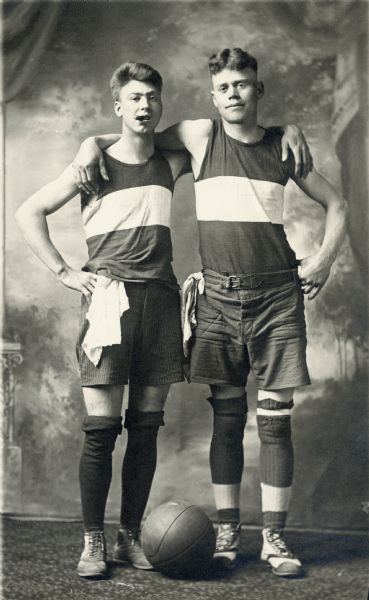 Photographic postcard of a studio portrait of two brothers in basketball uniforms with their arms around each other's shoulders. They are standing in front of a painted backdrop. The man on the left, with a cigar in his mouth, is unidentified, while the man on the right is Rudolph Faust.